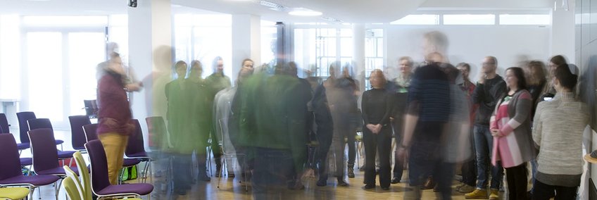 A group of around 30 people are standing in a circle in a seminar room. Most of the people are artistically blurred.