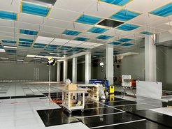 Spacious construction site featuring suspended ceiling panels partially covered with blue protective film, work equipment, lighting setups, and a ladder in the background.