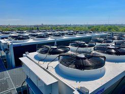 Industrial cooling systems with large fans on a building roof. In the background, a cityscape stretches out under a blue sky.