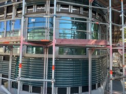 Rundes Gebäude mit Keramikfassade und Fenstern, umgeben von einem Gerüst für Bauarbeiten, reflektiert die Umgebung.