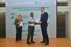 Im Fokus des Bildes stehen zwei Männer; der eine überreicht dem anderen ein Päckchen. Im Hintergrund sieht man eine weitere Person und ein großes Plakat mit dem Titel "20th Anniversary of Max-Planck India Partner Groups".