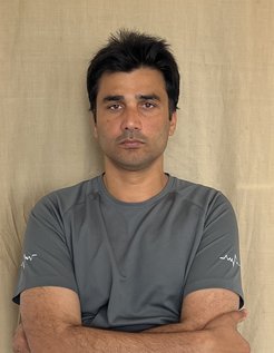 Person in gray athletic shirt with white graphic on sleeves, arms crossed, standing against a beige fabric backdrop.