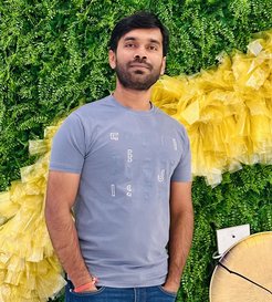 Person in casual attire, grey shirt with abstract text design, standing against a backdrop of lush greenery and vibrant yellow accents, next to a circular object.