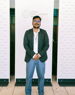 A persont in formal dress, including a grey blazer and blue trousers, poses before a Lindau Nobel Laureate Meeting banner.