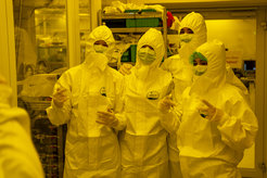 Four people wear full protective suits and masks in a laboratory room with scientific equipment and shelves.
